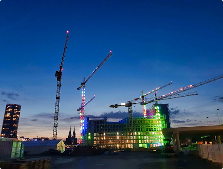 A construction site at dusk, featuring several cranes adorned with colorful lights in red, green, and white. Buildings are visible in the background, with a deep blue sky above.